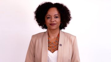 Mulher negra, de cabelos pretos, usando jaqueta bege e camiseta branca