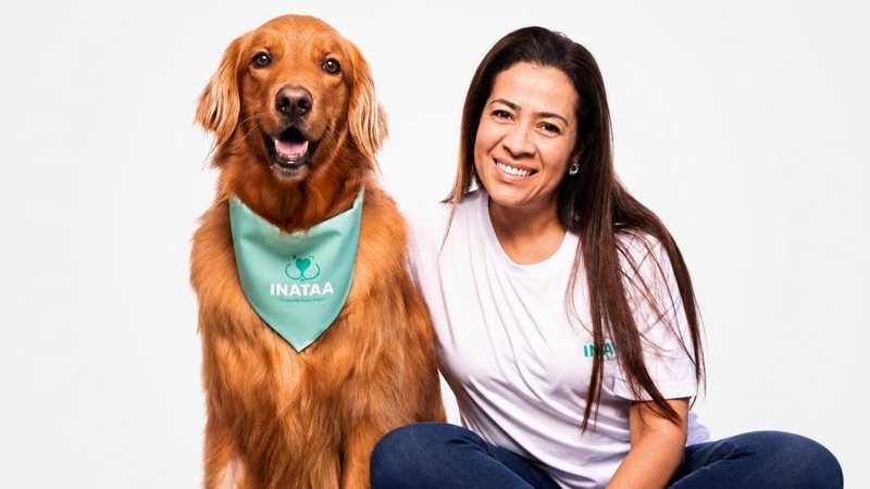 Foto de Gabriella, uma mulher branca de cabelos castanhos e longos com camiseta e jeans, e Zoff, um golden retriever usando uma bandana verde