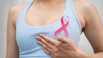 Mulher com camiseta azul e fita rosa da campanha Outubro Rosa apalpa o seio.