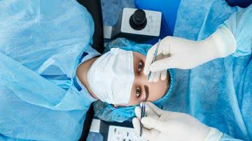 Paciente mulher paramentada e duas mãos com luvas cirúrgicas segurando instrumentos cirúrgicos sobre seus olhos