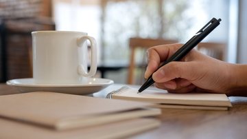 mesa com cadernos, xícara e pires e uma mão segurando uma caneta e escrevendo