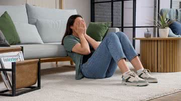 Jovem sentada no chão, encostada no sofá, segura o rosto com as mãos parecendo exausta