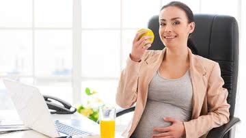 Mulher grávida come fruta e põe a mão na barriga em frente a computador