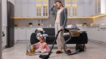 Mulher com expressão cansada segura aspirador e pó enquanto cuida de criança que brinca no chão