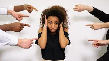 mulher ao centro com as mão nas orelhas e diversos dedos apontando para ela