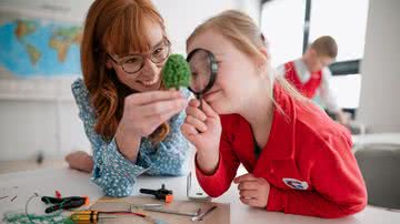 Criança atípica olha objeto com lente de aumento ao lado de professora